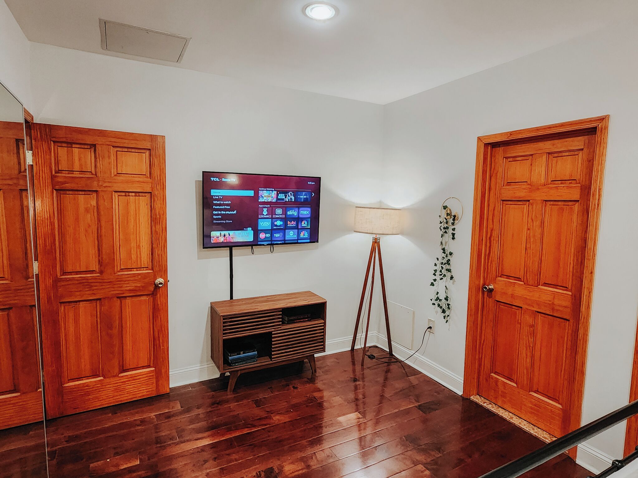A hallway with wooden doors, a flat-screen TV mounted on the wall, a floor lamp, and a wooden entertainment unit.