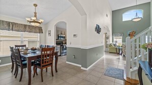 Une vaste salle à manger avec un plafond haut, une cuisine à l’arrière-plan et un salon avec un escalier.