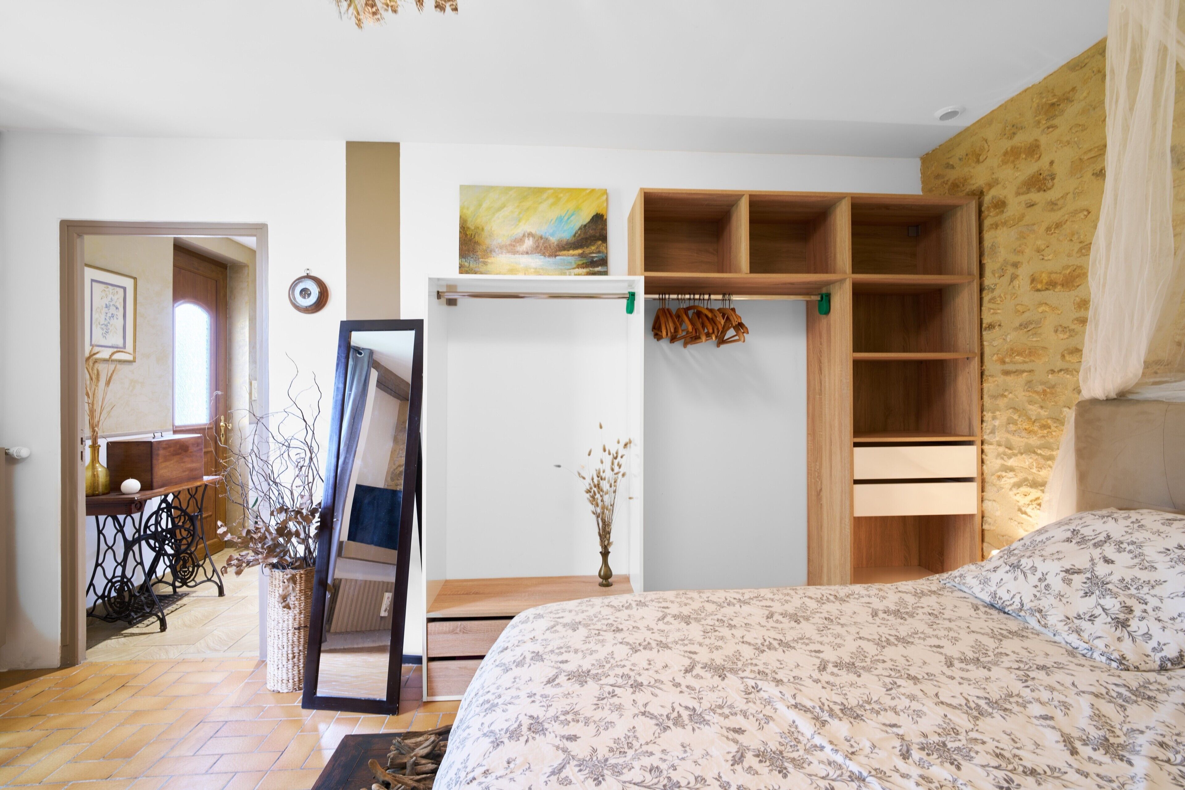 Une chambre à coucher avec un lit, une armoire en bois, un miroir, un tableau et un vase contenant des plantes séchées.