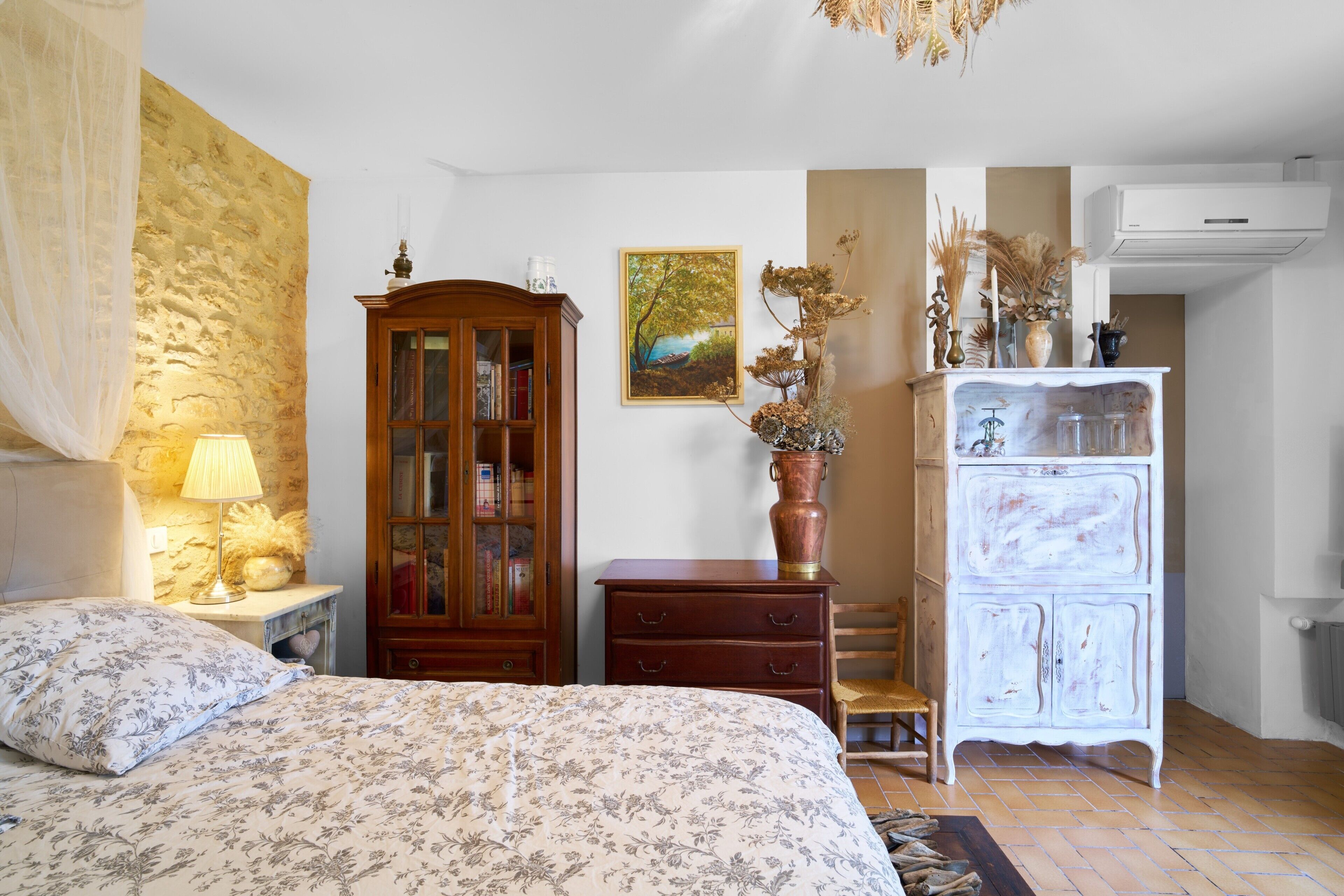 A bedroom with a bed, a wooden cabinet, a dresser, a lamp, and a painting on the wall.
