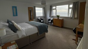 A hotel room with a bed, a desk, a chair, and a dresser. There are windows with curtains, a plant on the dresser, and a painting on the wall.
