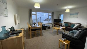 A living room with a sofa, a dining table with chairs, a TV, and a view of the sea.