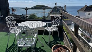 A balcony with a white metal table and chairs, overlooking a coastal view with a small island and buildings in the distance.