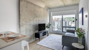 Ein modernes Wohnzimmer mit großem Fenster, einem Flachbildfernseher, einem grauen Sofa und einem Esstisch mit Tellern und Gläsern.