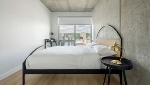 Ein modernes Schlafzimmer mit einem großen Bett, einem runden Tisch mit Lampe und Blick auf die Stadt durch große Fenster.