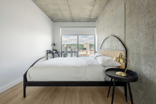 A modern bedroom with a large bed, a round table with a lamp, and a view of the city through large windows.