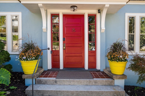 A red door with the number 15, flanked by two yellow flower pots on a set of steps leading to a blue house with white trim.