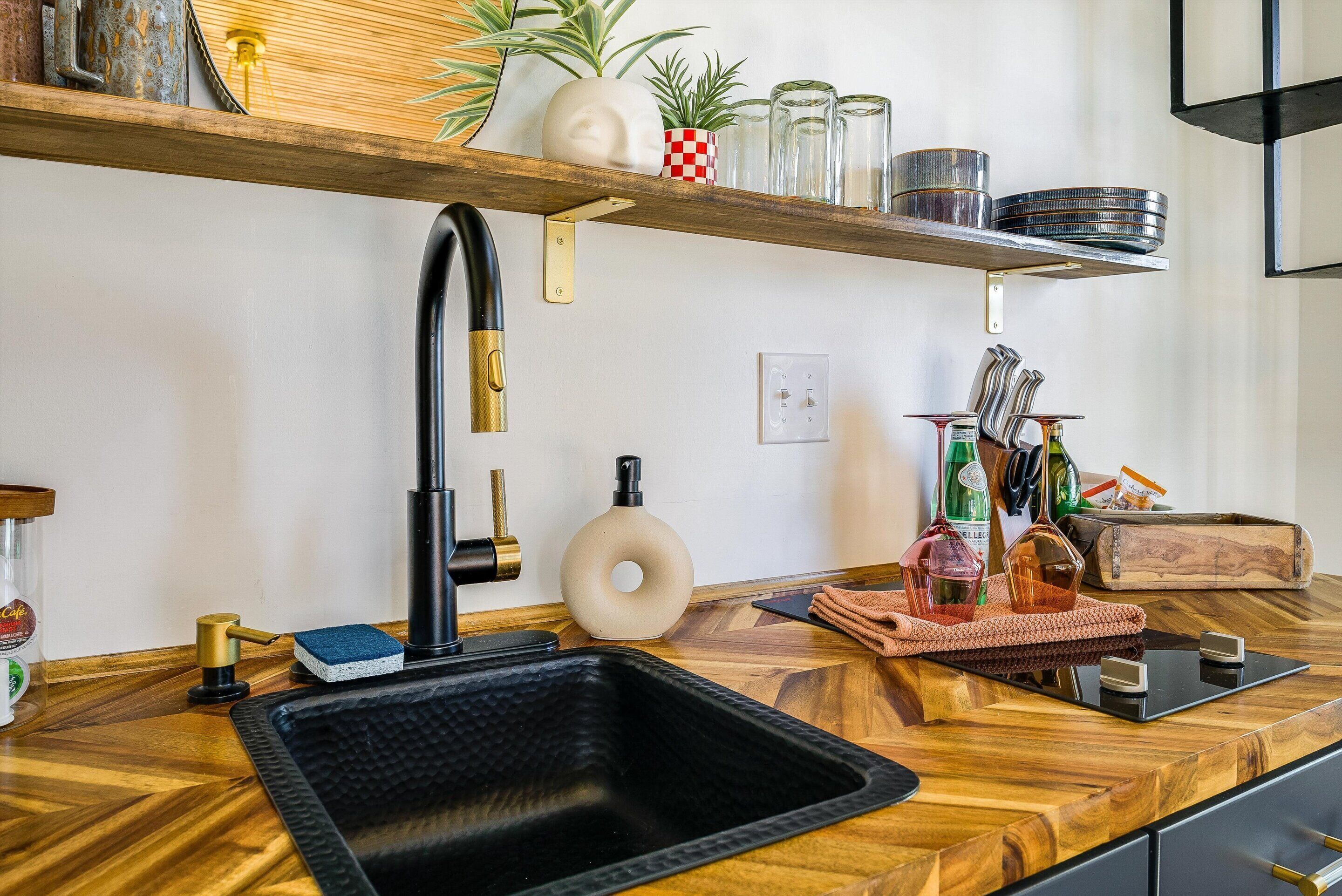 Une cuisine moderne avec un évier noir, un plan de travail en bois et une étagère au-dessus où sont rangés divers ustensiles de cuisine.