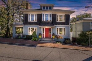 A two-story house with a red front door, white trim, and a small porch with potted plants.