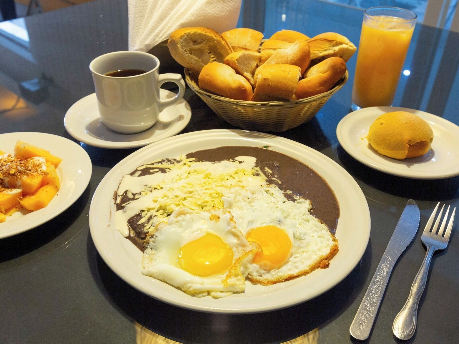 Une table mise pour le petit-déjeuner avec une assiette d’œufs, une tasse de café, un panier de pain et un verre de jus d’orange.