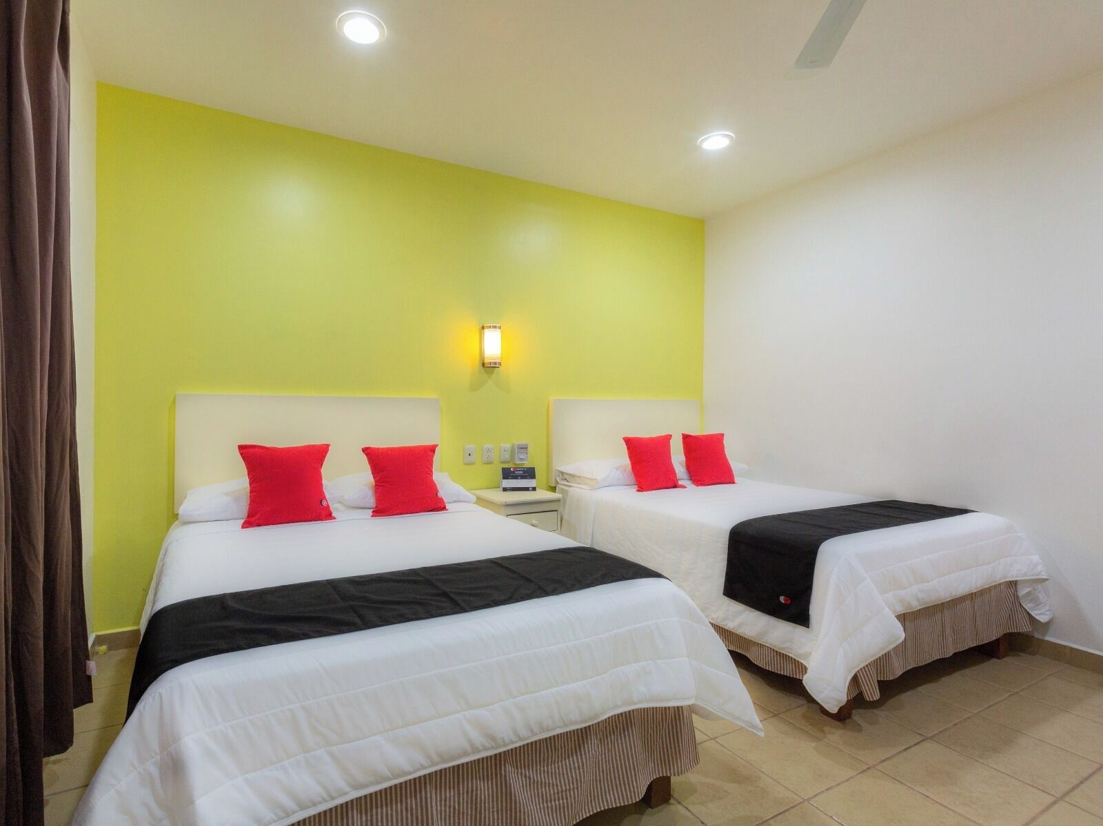 A hotel room with two beds, each with white bedding and red pillows, a nightstand, and a wall-mounted light.