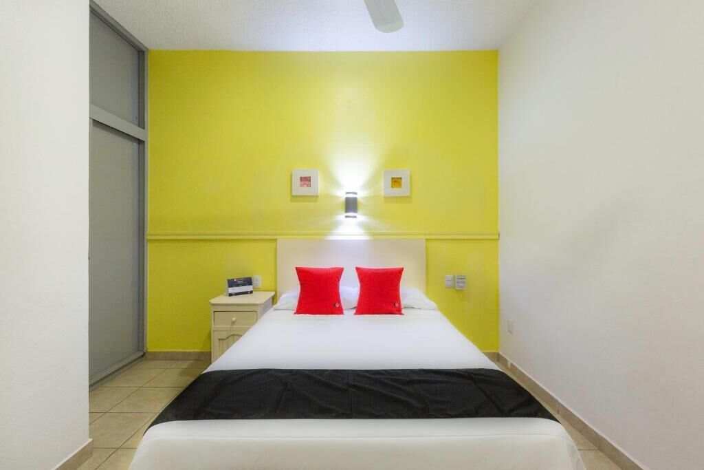 A small, brightly lit room with a bed, a nightstand, and a wall-mounted light fixture.