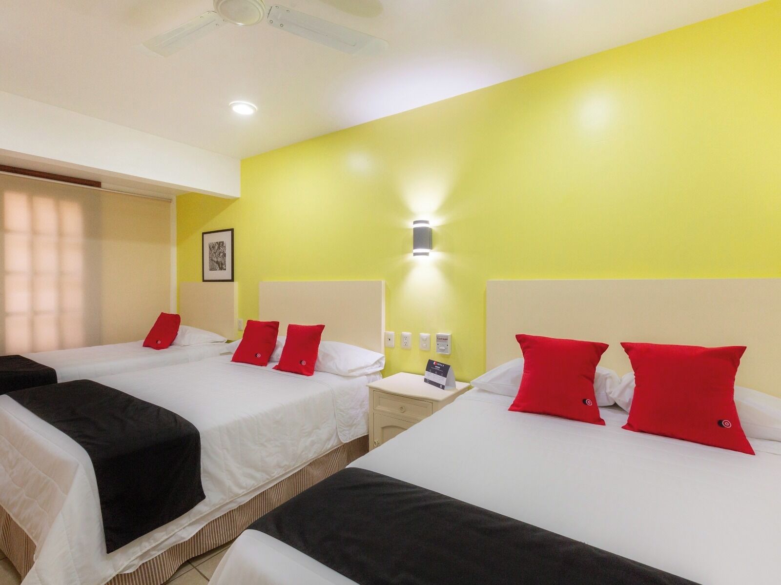 A hotel room with two beds, each with white bedding and red pillows. There is a bedside table with a lamp and a phone. The room has a yellow accent wall and a ceiling fan.