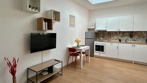 Una cocina moderna con armarios blancos, una zona de comedor con mesa y sillas, un televisor de pantalla plana montado en la pared y un tragaluz.