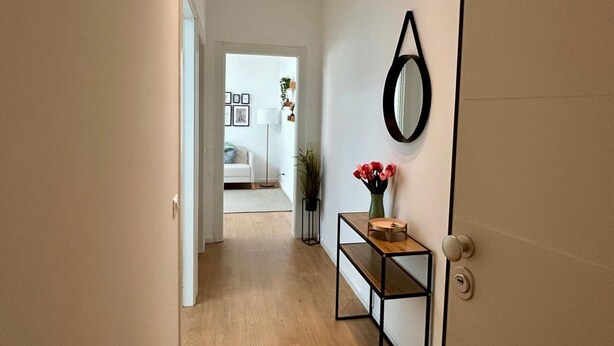 Un pasillo con piso de madera, una puerta con espejo redondo, una pequeña mesa con un florero y una puerta blanca que da a una habitación con cama y cuadros enmarcados.