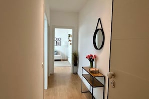 Un pasillo con piso de madera, una puerta con espejo redondo, una pequeña mesa con un florero y una puerta blanca que da a una habitación con cama y cuadros enmarcados.