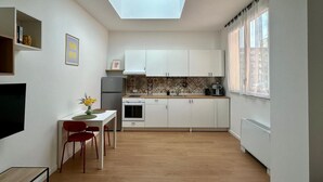 Cocina compacta con armarios blancos, tragaluz, zona de comedor con mesa y sillas, y ventana con cortinas traslúcidas.