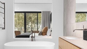 Uma casa de banho moderna com uma banheira independente, um espelho grande e vista para uma varanda com uma cama e uma cadeira.