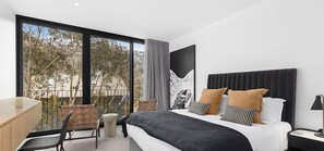 Une chambre d’hôtel moderne dotée d’un grand lit, d’un bureau et d’un balcon offrant une vue sur les arbres et les bâtiments.