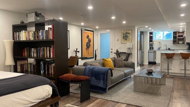 A modern living room with a sofa, a bookshelf, a dining area with a kitchen in the background, and a bed.
