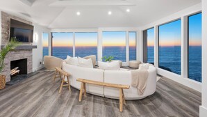 A modern living room with a large white sectional sofa, a wooden bench, and a fireplace. It features large windows with a view of the ocean and a skylight above. - Distinguished Oceanfront Point Estate with Iconic Beach + Mountain Vistas (Montara)