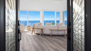 A modern living room with a white sofa, wooden benches, and a view of the ocean through large windows. - Distinguished Oceanfront Point Estate with Iconic Beach + Mountain Vistas (Montara)