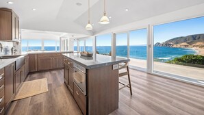A modern kitchen with a large island, wooden cabinets, and a view of the ocean. - Distinguished Oceanfront Point Estate with Iconic Beach + Mountain Vistas (Montara)