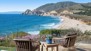 A coastal view with a beach, chairs, and a table with a plant. - Distinguished Oceanfront Point Estate with Iconic Beach + Mountain Vistas (Montara)