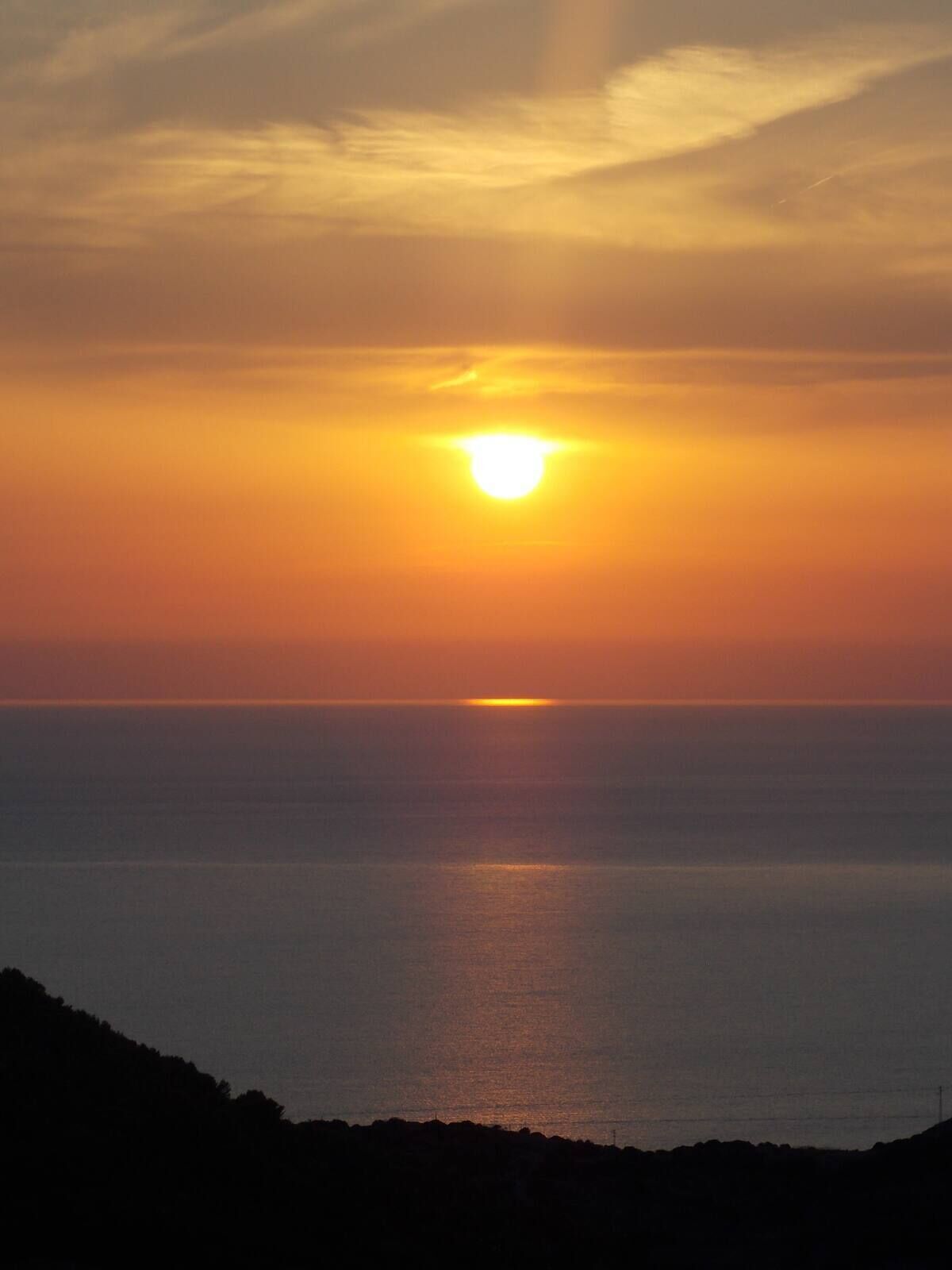 A sunset over a calm sea with a silhouette of a hilly landscape in the foreground.