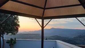 Pátio externo com mesa posta para refeições, com vista para uma paisagem montanhosa durante o pôr do sol.