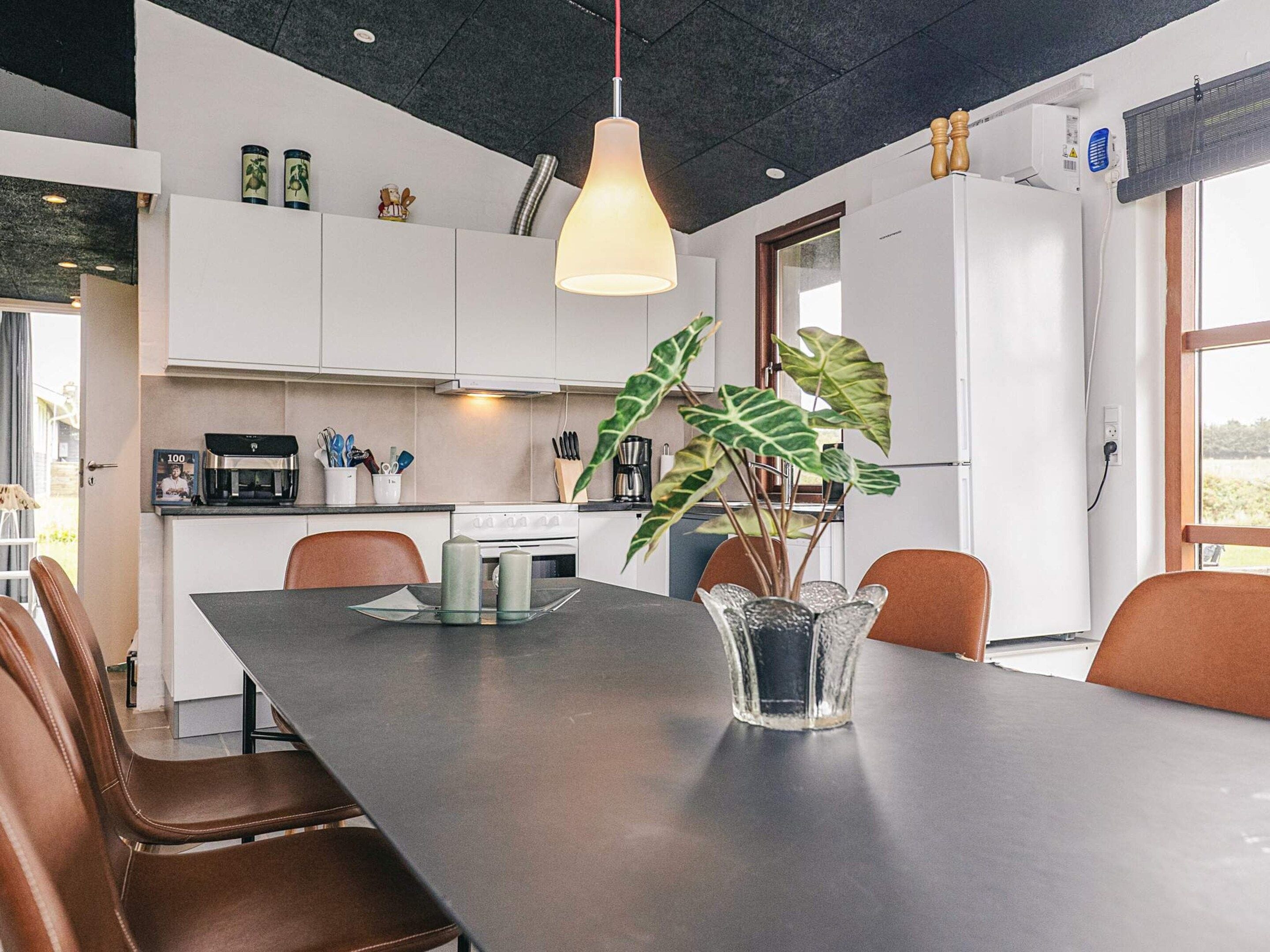 A modern kitchen with white cabinets, a dining table with chairs, a pendant light, and a potted plant.