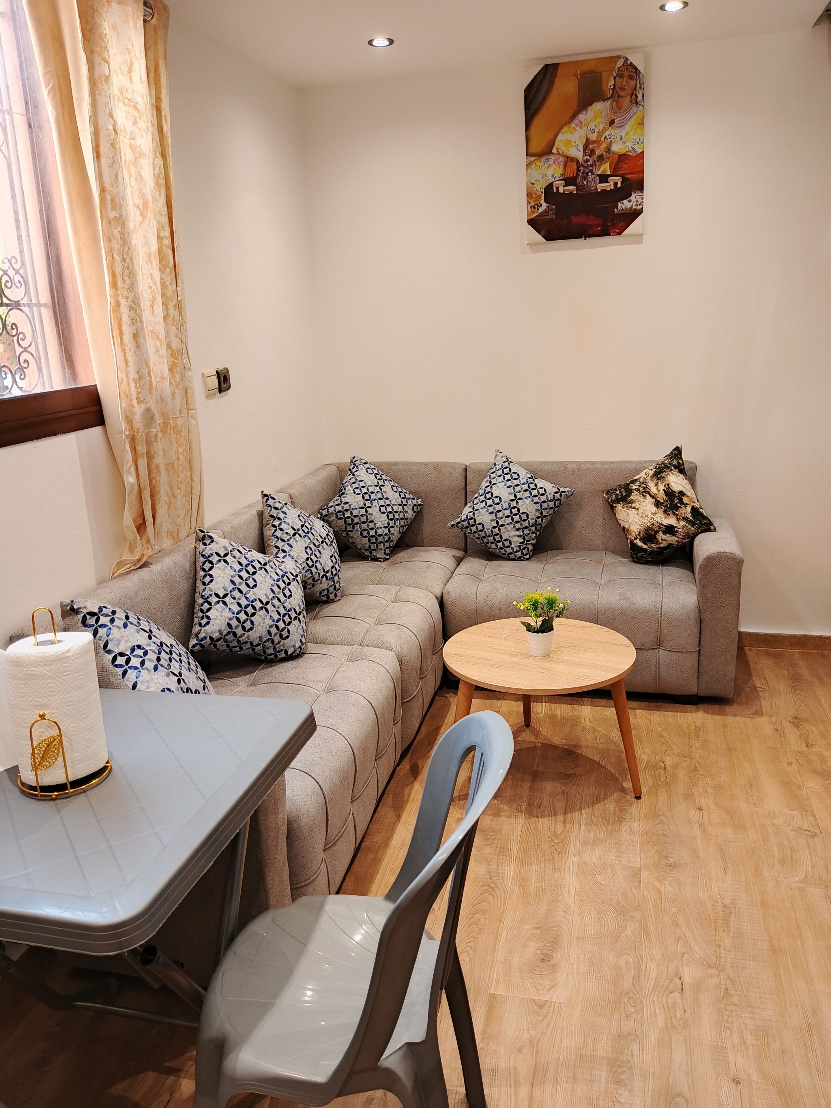 Un salon moderne avec un canapé d’angle, des coussins à motifs, une table basse ronde en bois, une table à manger avec une chaise et une plante en pot.