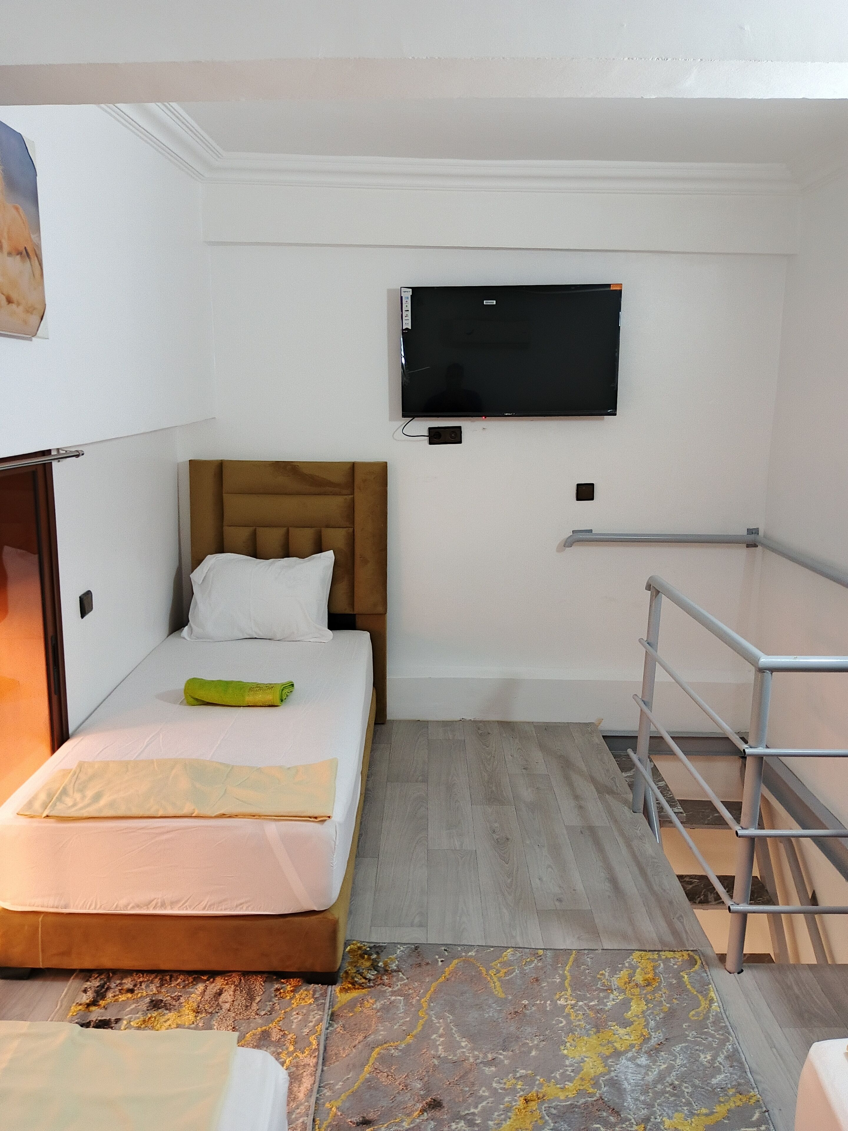 Une petite pièce bien éclairée, avec un lit, un téléviseur à écran plat fixé au mur, un plancher en bois et une rambarde en métal.
