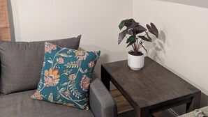 A living room with a grey sofa, a colorful floral pillow, a framed picture of a lion in a bathtub, a small wooden table with a potted plant, and a dark wooden floor. - Perfectly Located 1 Bedroom, newly remodeled  (Augusta)