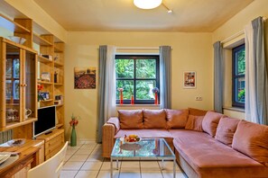Ein Wohnzimmer mit einer Couch, einem Glastisch, einem Fernseher, Holzregalen und einem Fenster mit Vorhängen.