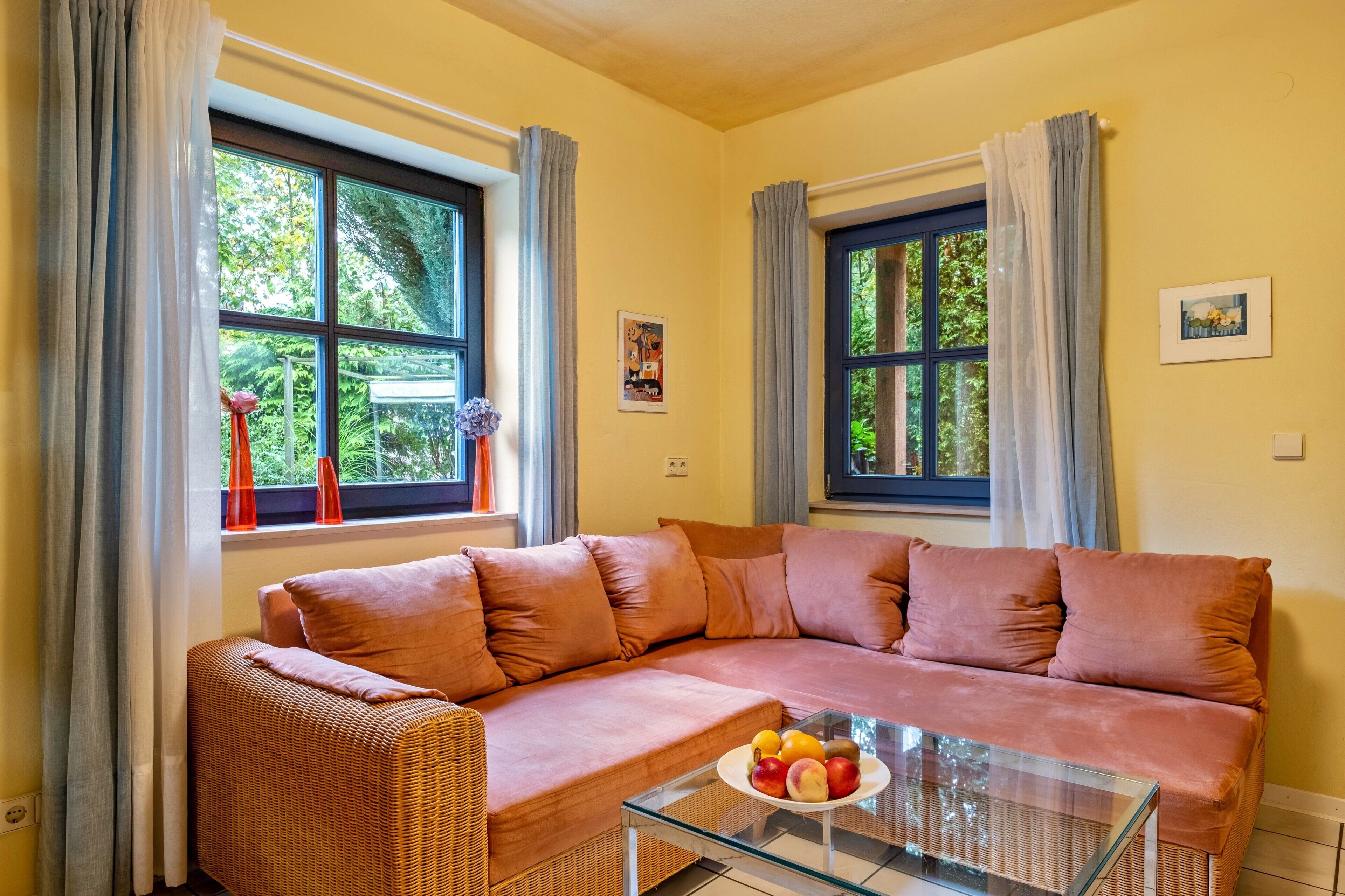Uma sala de estar com um sofá, uma mesa de centro de vidro com frutas e duas janelas com cortinas.