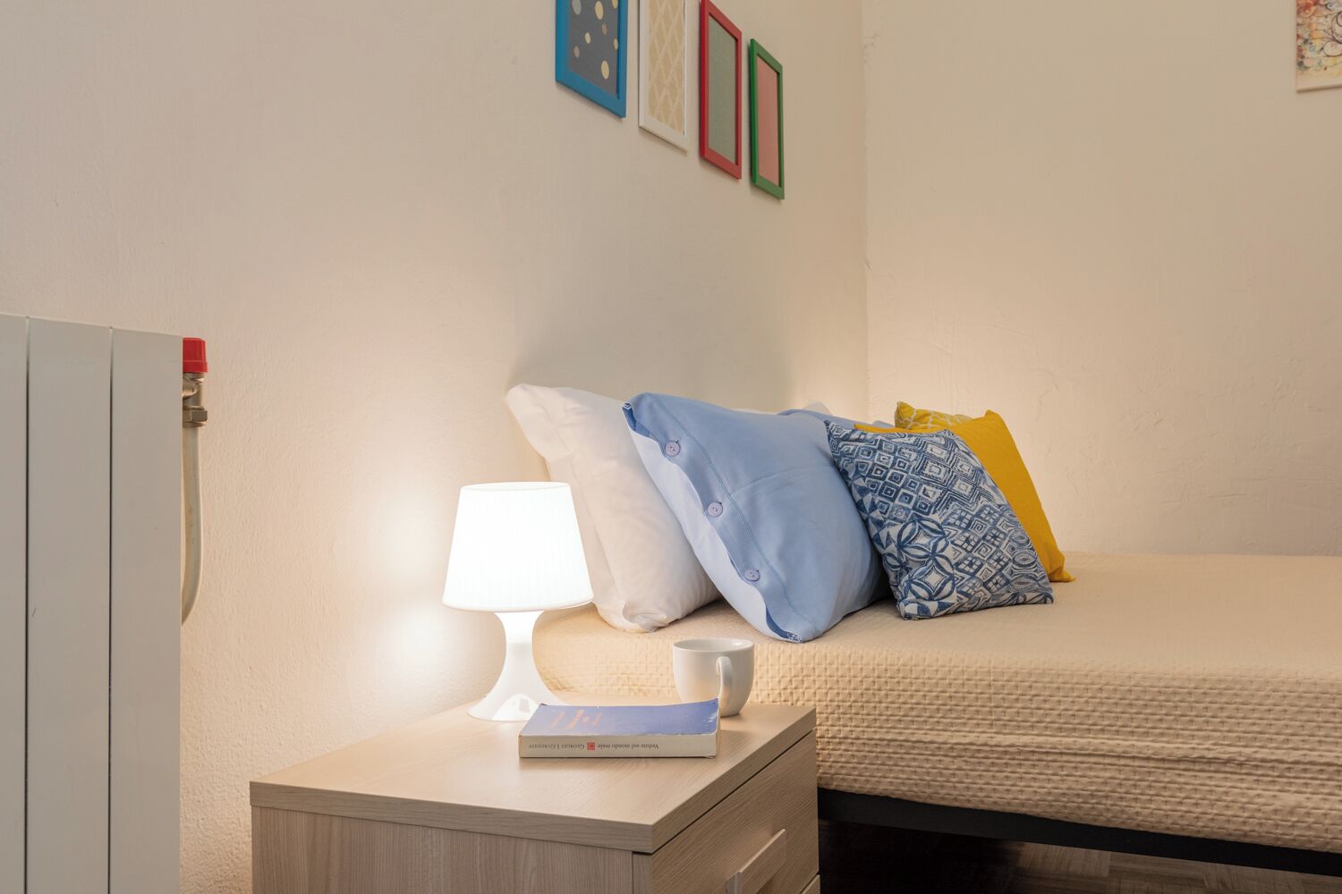 A neatly made bed with pillows, a bedside table with a lamp and a book, and a wall with framed pictures.