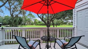 A covered outdoor seating area with two chairs and a table, a red umbrella, and a view of a green lawn and trees. - Dog friendly 1st floor  King bd in Ocean Ridge (Edisto Island)