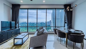 A modern living room with a sofa, coffee table, dining table, and chairs, large windows with a city view, and a ceiling fan. - Opus Residences by Wakely Kuala Lumpur (Kuala Lumpur)