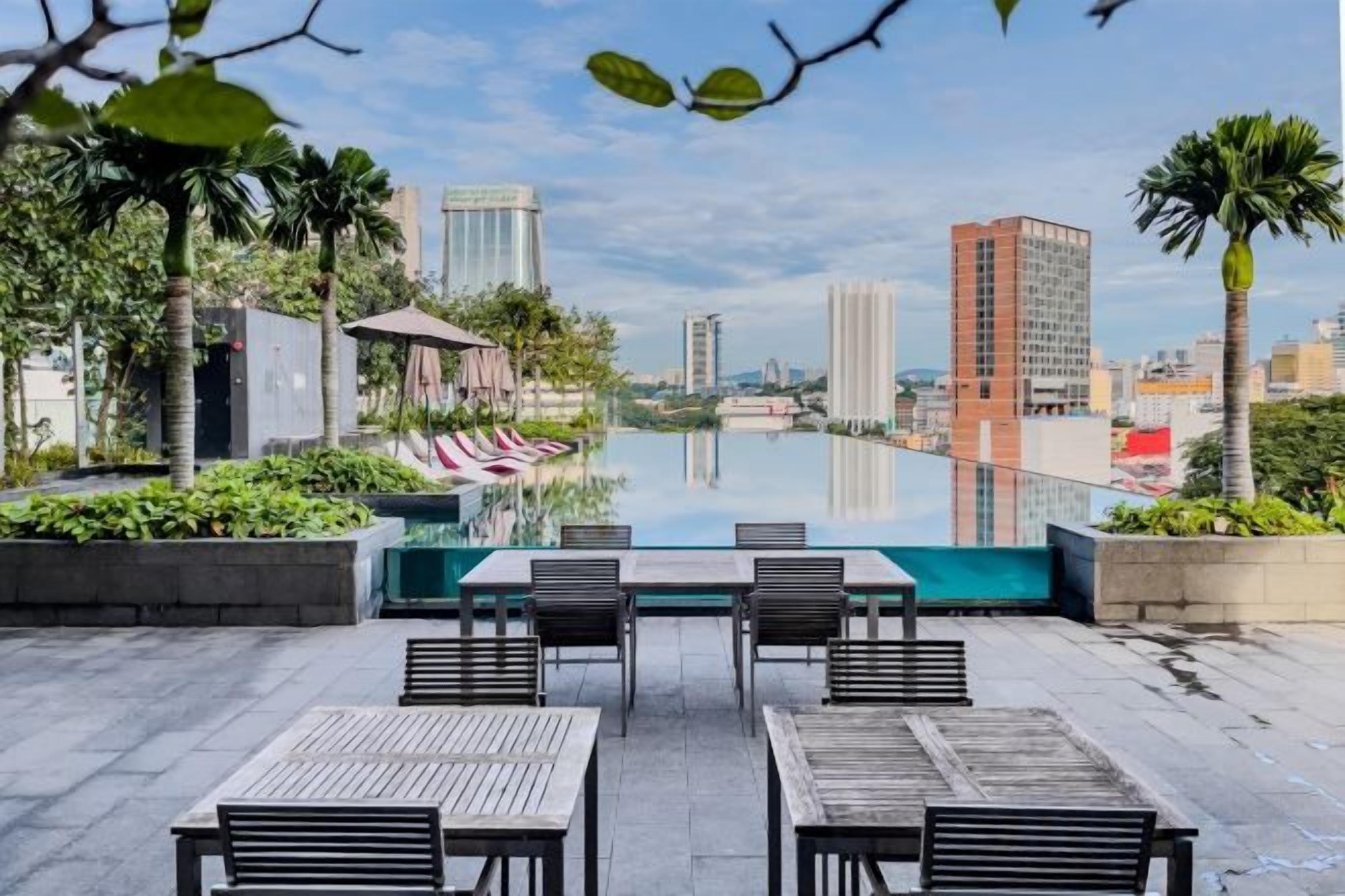 A rooftop pool area with tables and chairs, surrounded by greenery and city buildings.