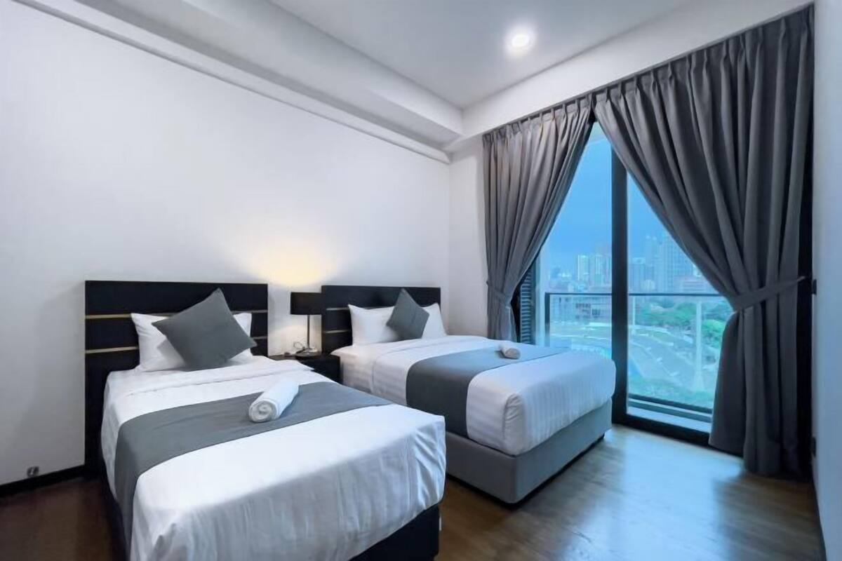 Habitación de hotel con dos camas, una mesita de noche, una lámpara y un ventanal con cortinas.