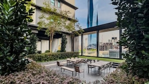 An outdoor seating area with tables and chairs, surrounded by greenery and large windows offering views of the cityscape. - Opus Residences by Wakely Kuala Lumpur (Kuala Lumpur)