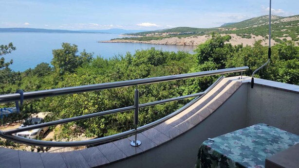 Un balcon doté d’une balustrade incurvée, offrant une vue sur un paysage côtier avec une étendue d’eau et des collines lointaines.