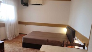 Une petite chambre avec un lit simple, une tête de lit en bois, un téléviseur fixé au mur et une fenêtre avec des rideaux.