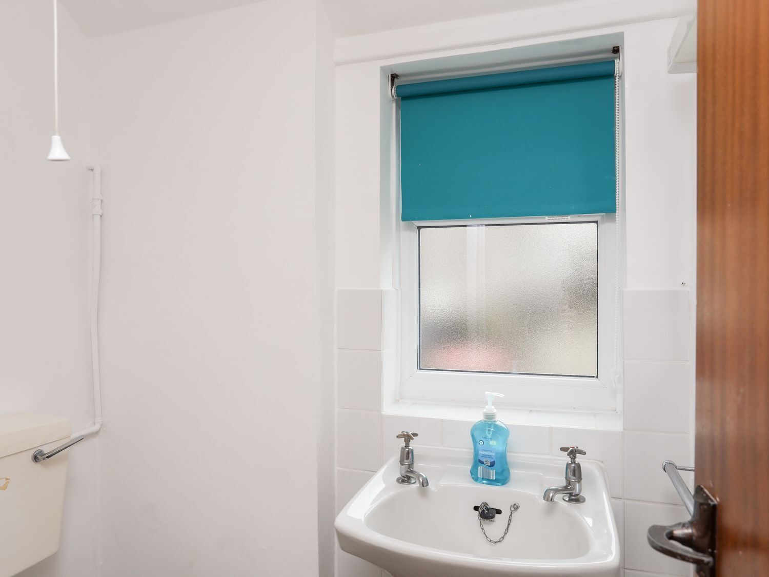 Un baño con un lavamanos blanco, un dispensador de jabón azul y un ventanal con persiana verde azulada.