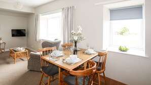 A dining area with a wooden table set for four, surrounded by chairs. There is a television mounted on the wall, a window with curtains, and a small side table with a basket. - Ceiriad - 9 Ty'n Don (Pwllheli)