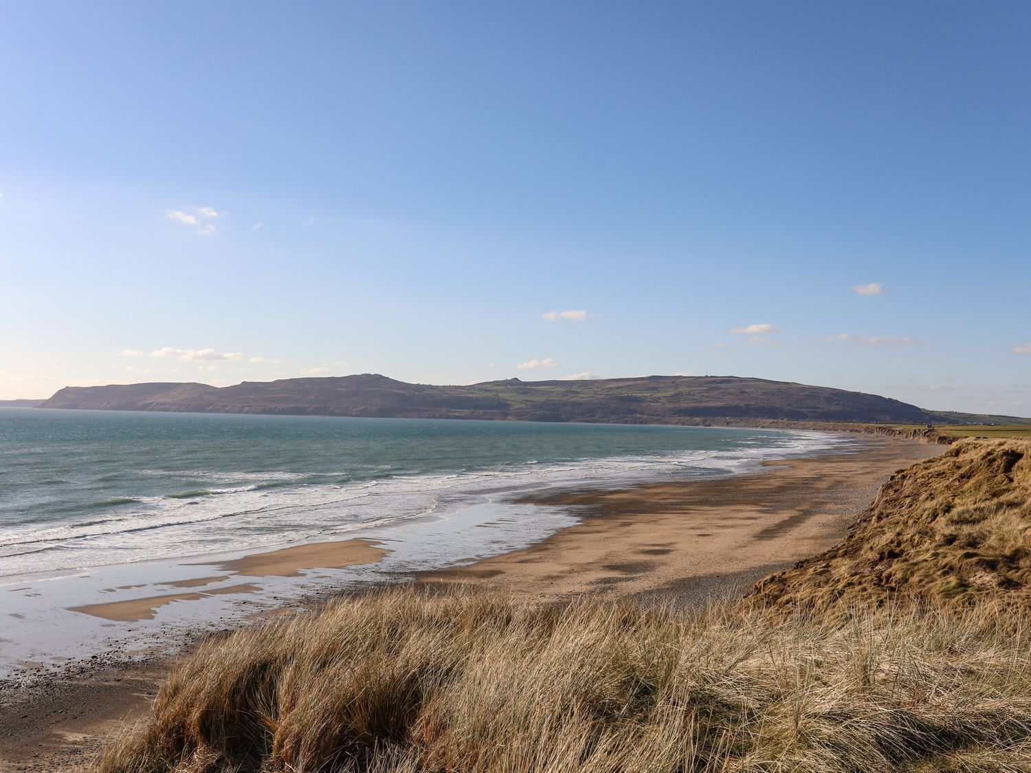 Un paysage côtier avec une plage de sable, des dunes herbeuses et un ciel bleu dégagé.