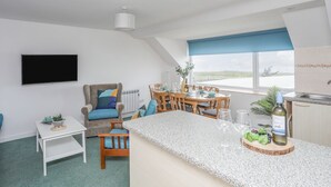 A modern living room with a TV, a dining area with a table set for two, and a kitchen with a window view. - Ysgo - 6 Ty'n Don (Pwllheli)