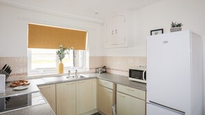 Una cocina moderna con armarios de madera clara, un refrigerador blanco, un microondas y una ventana con persiana color beige.