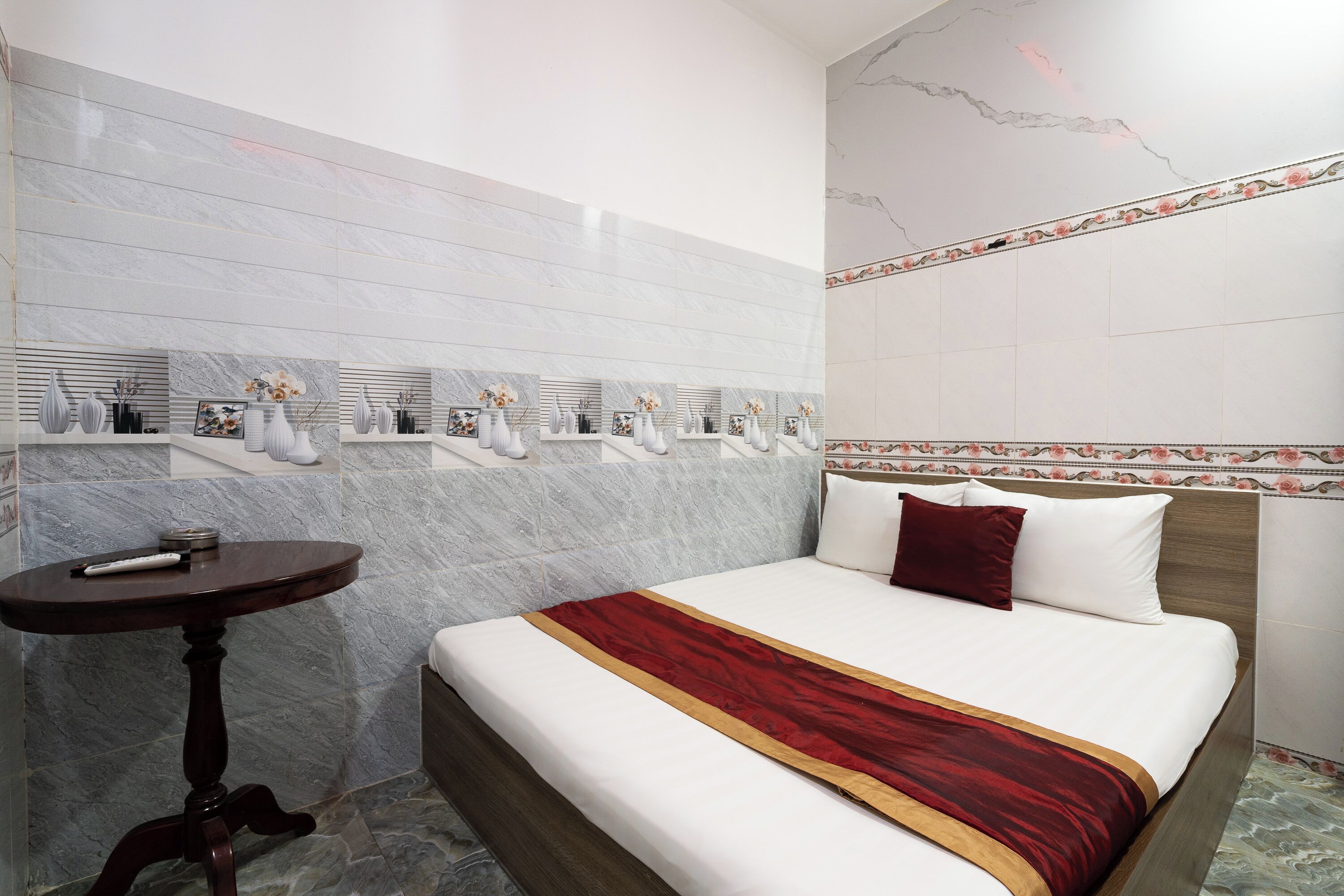 A hotel room with a marble-tiled wall, a bed with white bedding and a red and gold throw, a small round table, and a decorative shelf with vases and a picture frame.